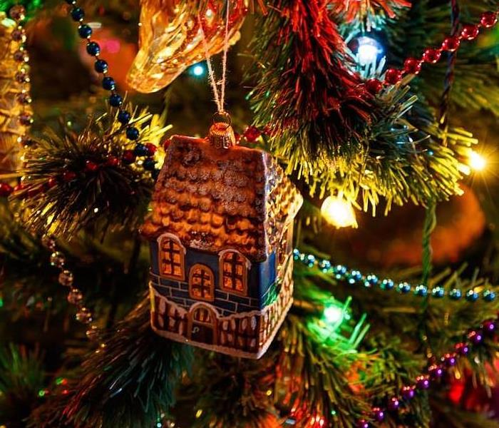 A Christmas tree ornament that looks like a house hangs on a bow amidst lights, beads, and other ornaments. 
